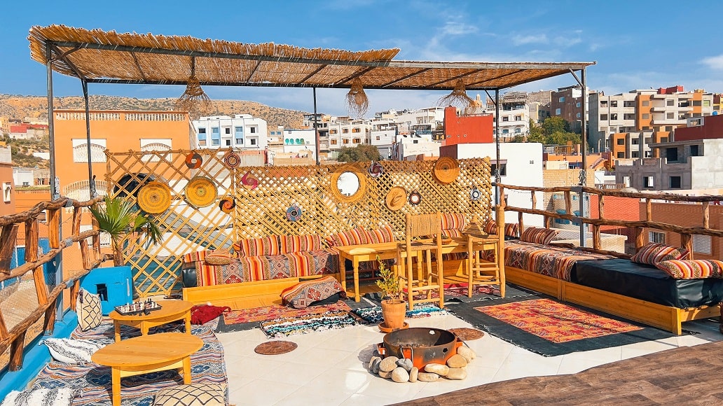 Terrasse rooftop ombragée du riad avec banquettes berbères, au-dessus du village