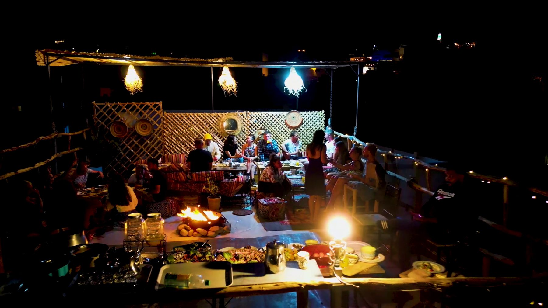 Long communal dinner table on the rooftop, guests eating together by lantern light